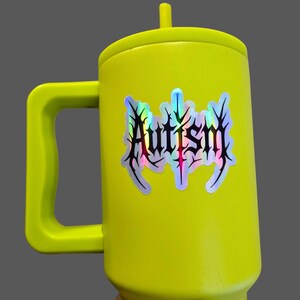 May include: A bright yellow mug with a handle and a lid. The mug features a holographic sticker with the word "Autism" in a stylized, gothic font. The mug is held against a gray background.