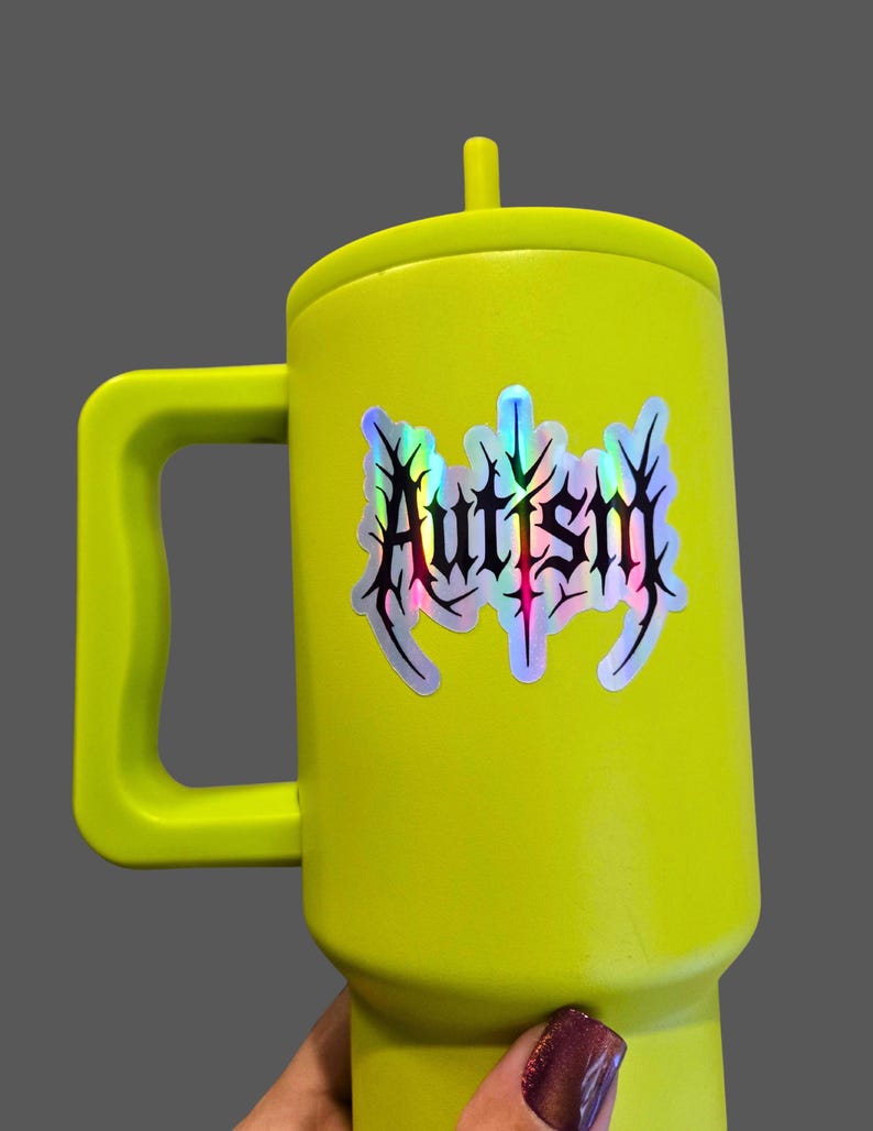 May include: A bright yellow mug with a handle and a lid. The mug features a holographic sticker with the word "Autism" in a stylized, gothic font. The mug is held against a gray background.