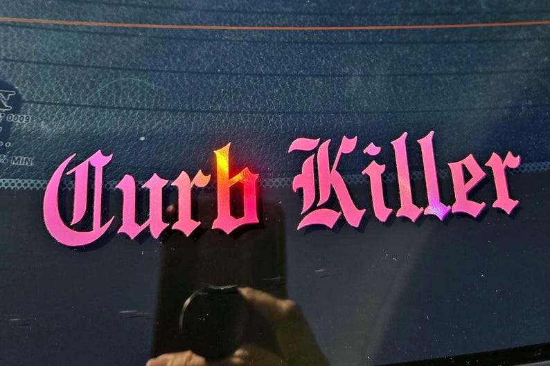 May include: A pink vinyl decal applied to a dark glass surface, featuring the text "Curb Killer" written in a stylized gothic font. The lettering displays a vibrant pink hue with a subtle gradient effect and a slight reflective quality.