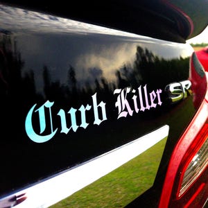 May include: A black car bumper featuring a decal with the text Curb Killer in a gothic font with a light blue to pink gradient. To the right of the text is a silver emblem with the letters SR. The surface is glossy and reflects the sky and trees.