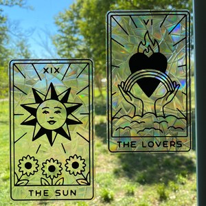Pode incluir: Dois cart&otilde;es de tar&ocirc; iridescentes com um efeito de vitral. O cart&atilde;o &agrave; esquerda &eacute; O Sol, cart&atilde;o n&uacute;mero XIX, com um sol com rosto e tr&ecirc;s girass&oacute;is. O cart&atilde;o &agrave; direita &eacute; Os Amantes, com duas m&atilde;os segurando um cora&ccedil;&atilde;o com uma chama e um arco-&iacute;ris.