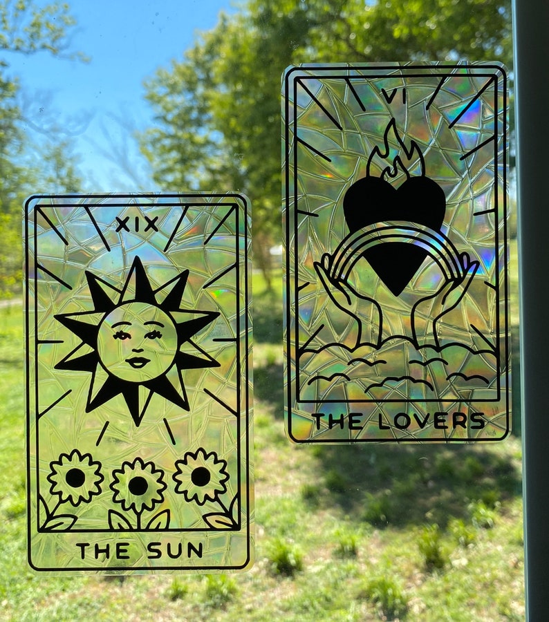 Pode incluir: Dois cart&otilde;es de tar&ocirc; iridescentes com um efeito de vitral. O cart&atilde;o &agrave; esquerda &eacute; O Sol, cart&atilde;o n&uacute;mero XIX, com um sol com rosto e tr&ecirc;s girass&oacute;is. O cart&atilde;o &agrave; direita &eacute; Os Amantes, com duas m&atilde;os segurando um cora&ccedil;&atilde;o com uma chama e um arco-&iacute;ris.