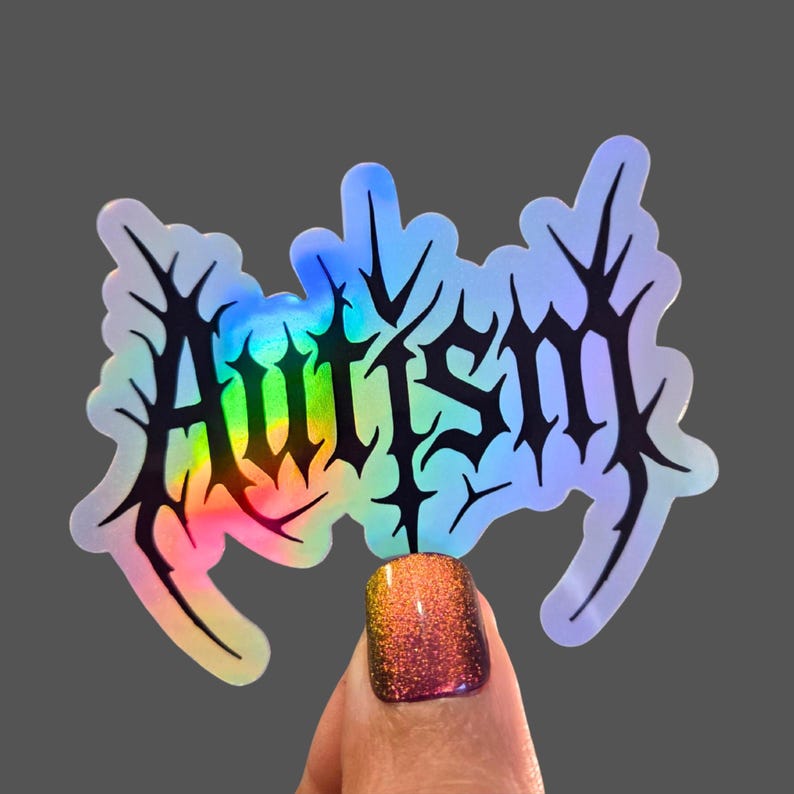 May include: Holographic sticker with the word "Autism" in a black, gothic font. The sticker has a rainbow gradient effect and is held up by a person's finger with a glittery nail.