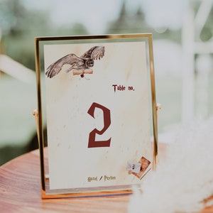 May include: A gold-framed table number card with a watercolor illustration of an owl carrying a scroll. The card has the number "2" in red and the text "Table no." and "Hazel / Peyton".