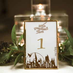 Puede incluir: Una tarjeta de número de mesa con marco dorado y fondo blanco. La tarjeta presenta una silueta marrón del castillo de Hogwarts, una pancarta que dice "Número de mesa" y el número "1". La esquina inferior derecha de la tarjeta dice "Haley & Peter".