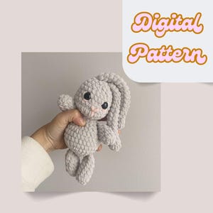 Puede incluir: Un conejito de ganchillo gris claro con grandes ojos negros y detalles rosas. El conejito se sostiene en una mano y tiene orejas largas y caídas. La imagen incluye el texto "Digital Pattern" en una fuente decorativa.
