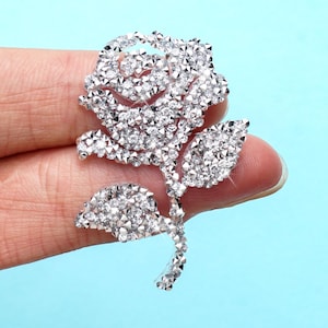 Peut inclure: Une broche en forme de rose argentée, ornée de nombreux cristaux transparents. Le motif floral comprend une fleur de rose détaillée, deux feuilles et une tige. La broche est posée sur un fond bleu clair.