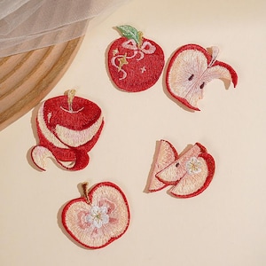 May include: Five embroidered apple-themed patches featuring a whole red apple with a bow, a bitten apple slice, a peeled apple, a cross-section of an apple showing seeds, and two overlapping apple slices, all rendered in shades of red, pink, and white.