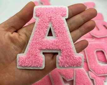May include: A pink, fuzzy letter "A" patch with a white border. The patch is held in a hand, with other letter patches visible in the background. The patches are likely for clothing or crafts.