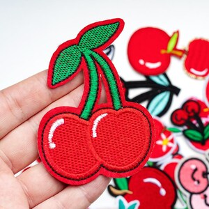 May include: Embroidered red cherry patch with green leaves and stems. The patch features a detailed design with white highlights on the cherries and a black outline. Other fruit patches are visible in the background.