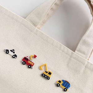 May include: A cream-colored tote bag with embroidered patches of toy vehicles: a police car, a fire truck, an excavator, and a dump truck. The patches are in red, yellow, black, white, and blue. The bag has two handles.