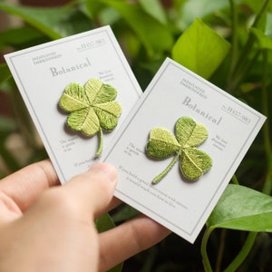May include: Two white cards with green embroidered four-leaf clover patches. The cards have the text "INFATUATED EMBROIDERED" and "Botanical" on them. The text "We love abundant nature." is also on the cards.
