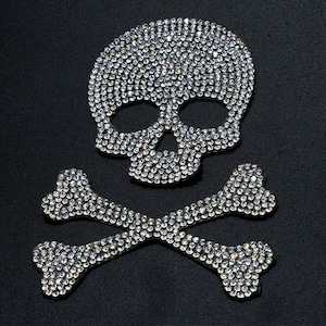 Op de afbeelding: Een zilveren schedel en gekruiste beenderen ontwerp gemaakt van strass. De schedel heeft een gedetailleerd gezicht met oogkassen, een neus en een mond. De gekruiste beenderen zijn achter de schedel geplaatst, met de uiteinden van de beenderen die in het midden kruisen.