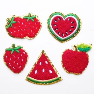 Può includere: Cinque toppe di frutta ricamate: fragole, una fetta di anguria a forma di cuore, una fragola singola, una fetta di anguria e una mela. Ogni toppa è rossa con dettagli verdi e bordi glitterati dorati.