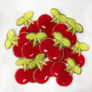 May include: A collection of red felt cherry patches with gold glitter accents and green felt stems. The cherries are arranged in a cluster, with some overlapping.