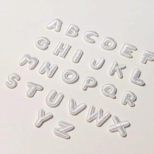 May include: A collection of white, embroidered alphabet letters arranged on a white surface. The letters are in a rounded, bubble-like font, and each letter is individually crafted with a textured, stitched appearance. The letters are from A to Z.