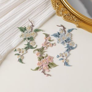 May include: Three embroidered floral branch patches featuring intricate stitching. One patch displays white flowers with green leaves, another shows pink blossoms with green foliage, and the third features light blue flowers with green leaves.