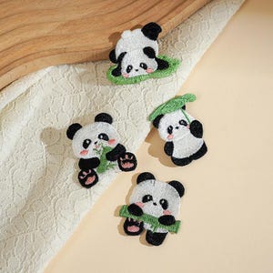 Può includere: Quattro toppe ricamate raffiguranti panda bianchi e neri. Un panda è sdraiato su una foglia verde, un altro tiene un gambo di bambù, un terzo è seduto sotto una grande foglia verde e il quarto tiene un segmento di bambù davanti al corpo.
