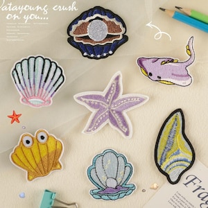 May include: A set of ten embroidered iron-on patches featuring various sea creatures and seashells. The patches are in shades of purple, blue, yellow, and pink. The patches include a starfish, a seashell, a clam, a stingray, and a seahorse.
