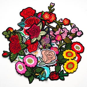 Op de afbeelding: Een verzameling geborduurde bloemenpatches in verschillende kleuren. De patches tonen rozen, madeliefjes en andere bloemen met groene bladeren en zwarte randen. Het kleurenpalet omvat rood, roze, geel en perzik.