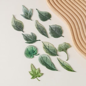 Könnte beinhalten: Eine Sammlung von zwölf gestickten Blatt-Aufnähern in verschiedenen Grüntönen. Das Set enthält diverse botanische Formen wie spitze, runde und gelappte Blätter, die jeweils mit detaillierten Fadenstickereien versehen sind, um natürliche Blattadern nachzuahmen.