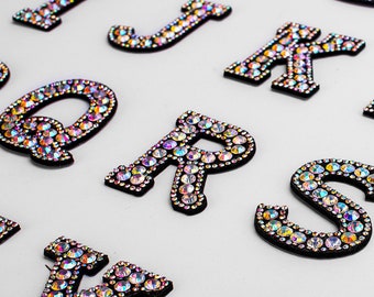 Può includere: Un set di lettere dell'alfabeto in feltro nero e ricoperte di strass iridescenti. Le lettere sono in maiuscolo e sono disposte in ordine casuale.