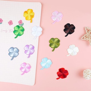 May include: A collection of embroidered four-leaf clover patches in a range of colours, such as yellow, green, white, blue, purple, pink, brown, and red. The patches are on a pink surface and a white notepad.