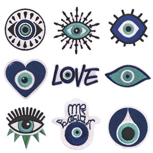 May include: Nine embroidered iron-on patches featuring the evil eye symbol in various styles. The patches are white with blue and green embroidery. The patches include a heart, a circle, a starburst, a hand, and an eye with eyelashes. The word "LOVE" is also included in one of the patches.