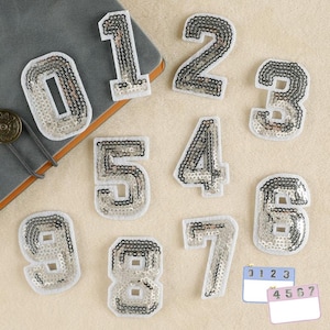 May include: Ten silver sequin iron-on patches in the shape of numbers 0 through 9. The patches are on a white background.