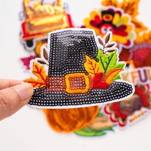 Può includere: Una toppa a forma di cappello da pellegrino nero con paillettes e fibbia gialla, decorata con foglie autunnali ricamate nei toni dell'arancione, rosso e verde. La toppa presenta spighe di grano marroni e un bordo bianco, a tema autunnale.