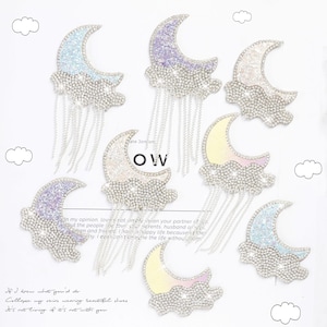 Può includere: Toppe a forma di luna crescente e nuvola decorate con catene d'argento pendenti. Le lune e le nuvole sono ricoperte di strass scintillanti in vari colori, tra cui blu, viola e iridescente. Le toppe sono probabilmente per abbigliamento o accessori.