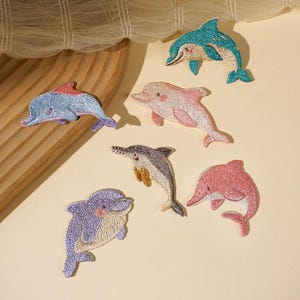 May include: A collection of six embroidered dolphin patches in various colors including teal, light pink, grey, purple, and coral. Each patch features detailed stitching to define the dolphin shape, fins, and facial features, presented as individual fabric appliques.