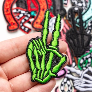 May include: A green embroidered patch in the shape of a skeleton hand making a peace sign. The patch has a black border and is surrounded by other patches with various designs.