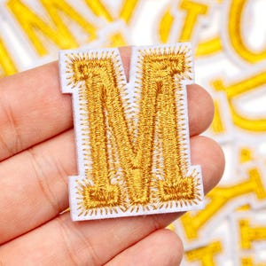 Pode incluir: Patch bordado com a letra "M" dourada sobre fundo branco. A letra é densamente bordada com fio dourado, criando uma aparência texturizada. O patch tem uma borda limpa e nítida e provavelmente é para aplicar em roupas ou acessórios.