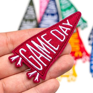 Può includere: Una toppa rossa a forma di gagliardetto con le parole "GAME DAY" ricamate in bianco. La toppa ha un bordo bianco e dettagli decorativi bianchi. Altre toppe a forma di gagliardetto in vari colori sono visibili sullo sfondo.