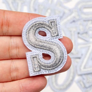 Può includere: Una toppa ricamata bianca a forma di lettera "S". La lettera è riempita con filo d'argento e delineata con un bordo argentato. La toppa è tenuta in mano da una persona.