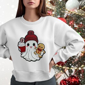 Op de afbeelding: Witte trui met een schattig cartoon-spookje dat een rode muts draagt en een kopje warme chocolademelk en een peperkoekman vasthoudt.