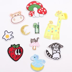 May include: A set of 10 iron-on embroidered patches featuring various cute cartoon characters and objects, including a strawberry, a mushroom, a giraffe, a cat, a hippo, a rocking horse, a moon, a fried egg, and a glass of juice.