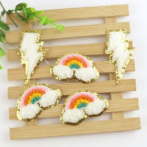 May include: Four iron-on patches with a rainbow, cloud, and lightning bolt design. The patches are white with gold glitter trim and colorful embroidery.
