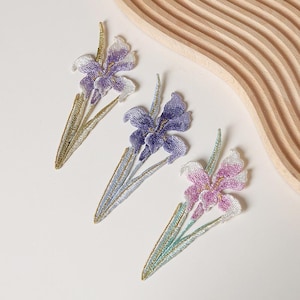 Op de afbeelding: Drie geborduurde irisbloem-patches met gedetailleerd stikwerk in tinten paars, wit en roze. Elke patch toont een lange groene stengel en bladeren met accenten van metallic gouddraad, wat zorgt voor ingewikkelde bloemige texturen en patronen.