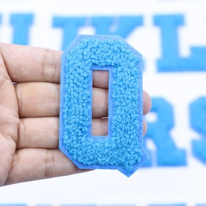 May include: A light blue felt letter O, with a slightly raised texture, is held in a hand. The letter is a common style used for sports team uniforms.