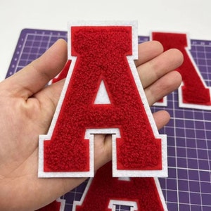 Può includere: Toppa rossa con la lettera "A" e bordo bianco. La lettera ha un aspetto strutturato e sfocato. Altre toppe con lettere rosse sono visibili sullo sfondo.