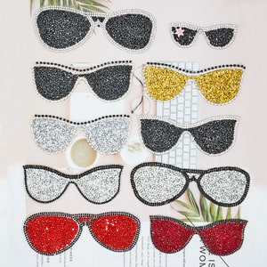 May include: An assortment of bedazzled sunglasses in various colors and styles. The sunglasses feature black, silver, gold, and red lenses, all encrusted with sparkling rhinestones. Some have a classic aviator shape, while others have a more modern, oversized design.