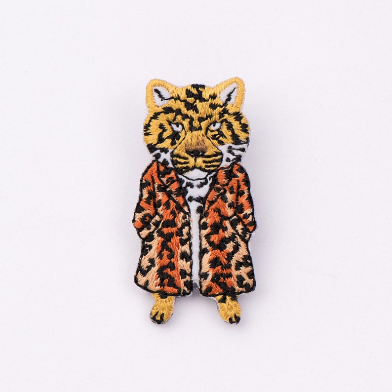 May include: Embroidered iron-on patch of a leopard wearing a leopard print coat. The patch is brown, orange, yellow, and white.
