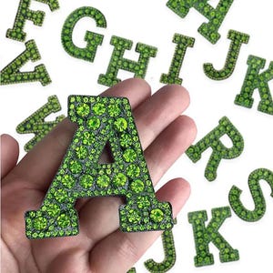 May include: Close-up of the letter "A" in a green rhinestone design, held in a hand. Other letters in the alphabet, also in green rhinestones, are scattered in the background. The letters have a dark border.