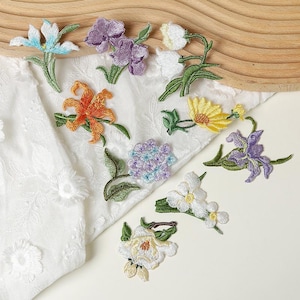 May include: A collection of assorted floral embroidered patches featuring various blooms including a blue and white flower, purple blossoms, white lily-of-the-valley, an orange lily, a yellow daisy, a purple iris, a hydrangea, and a white rose.