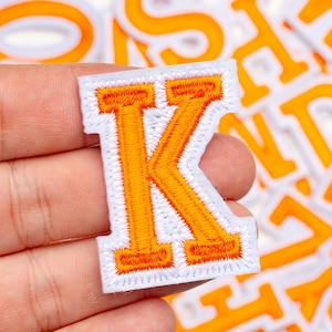 Può includere: Toppa ricamata con la lettera "K" con cuciture arancioni su sfondo bianco. La toppa ha un classico stile lettera universitaria. Altre toppe con lettere arancioni sono visibili sullo sfondo.