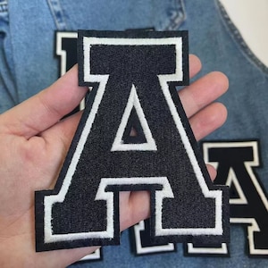 Pode incluir: Letra A de feltro preto com contorno branco, em uma fonte de estilo universitário. A letra é termocolante e pode ser usada para personalizar roupas, bolsas ou outros itens.