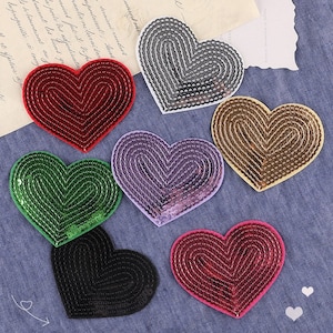 May include: Six heart-shaped patches with sequin embellishments in various colors, including red, green, black, silver, purple, and gold. The patches are arranged on a blue denim fabric background.