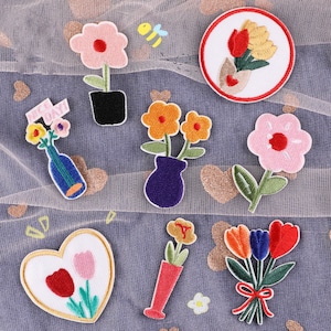 May include: Assortment of embroidered iron-on patches featuring floral designs, a bee, and a bouquet of flowers in a vase. The patches are on a white mesh background.